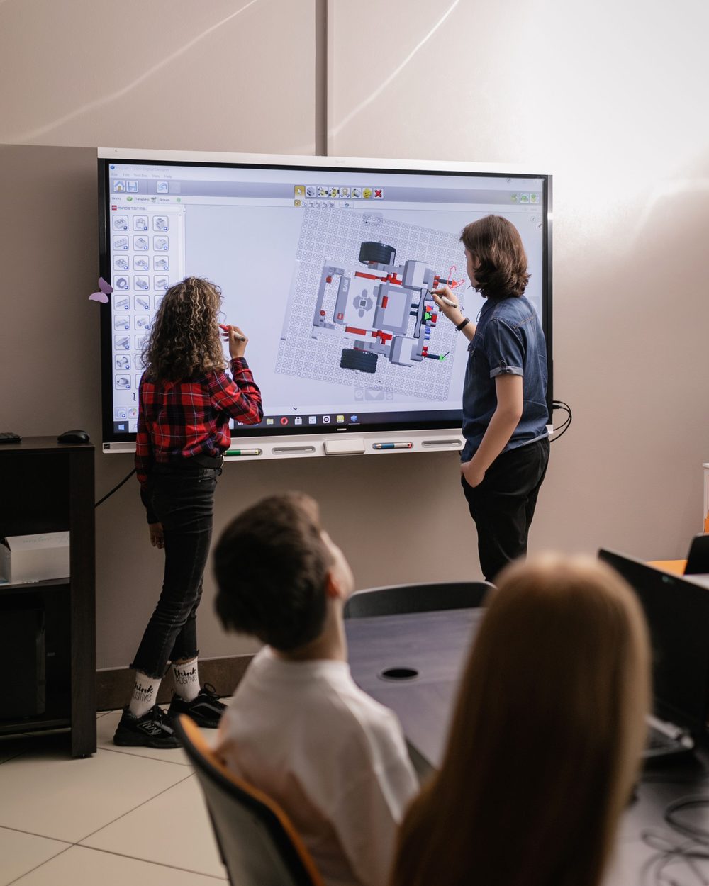 Two students at a smartboard building a robot design while classmates watch